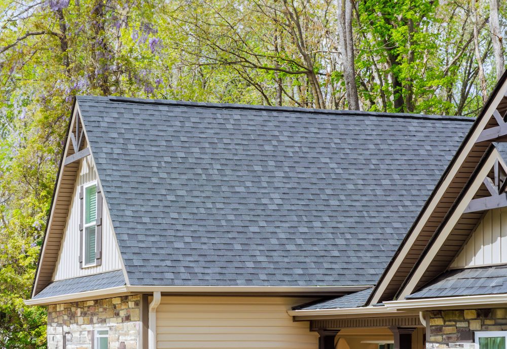 A close-up shot of a roofing contractor inspecting asphalt shingles on a house. The roof is set against a clear blue sky, showcasing freshly repaired shingles and emphasizing durability and quality craftsmanship.