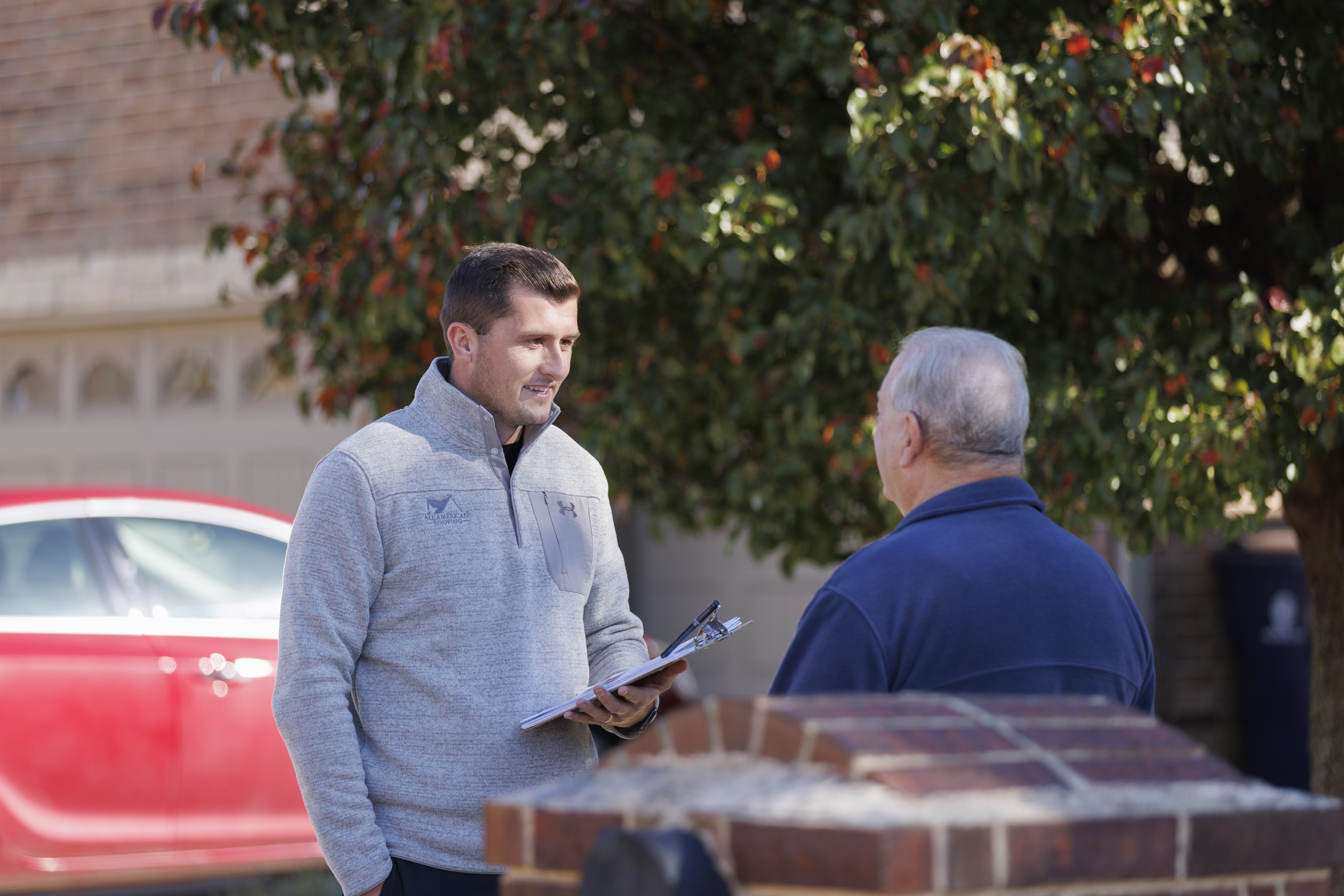 A roofing representative in a light gray quarter-zip pullover with the All American Roofing logo speaks with an older man in a navy shirt outside a suburban home. The representative holds a clipboard while standing near a red car and a brick mailbox, with a leafy tree in the background.