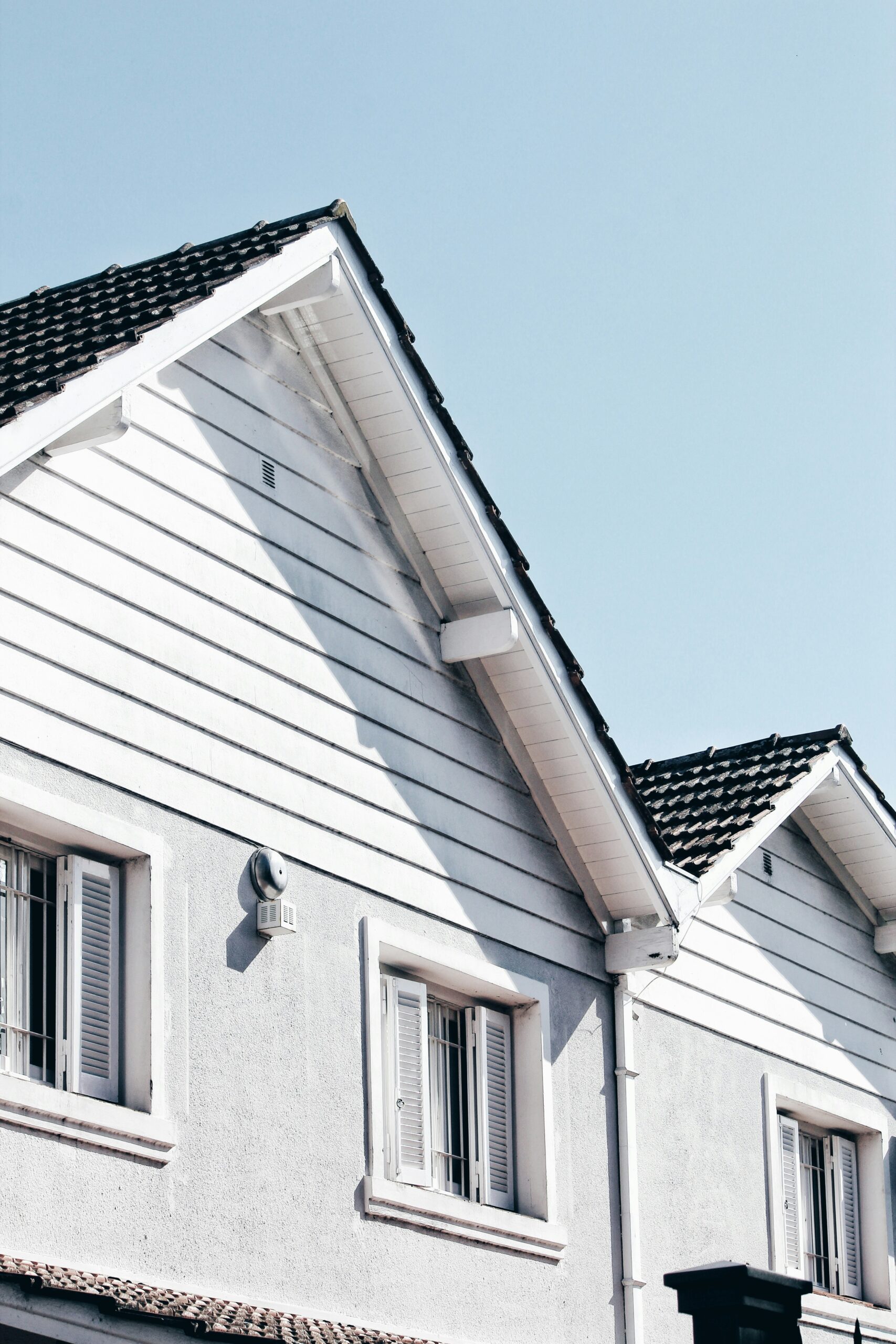 A white house with dark roof shingles and shuttered windows under a clear blue sky.