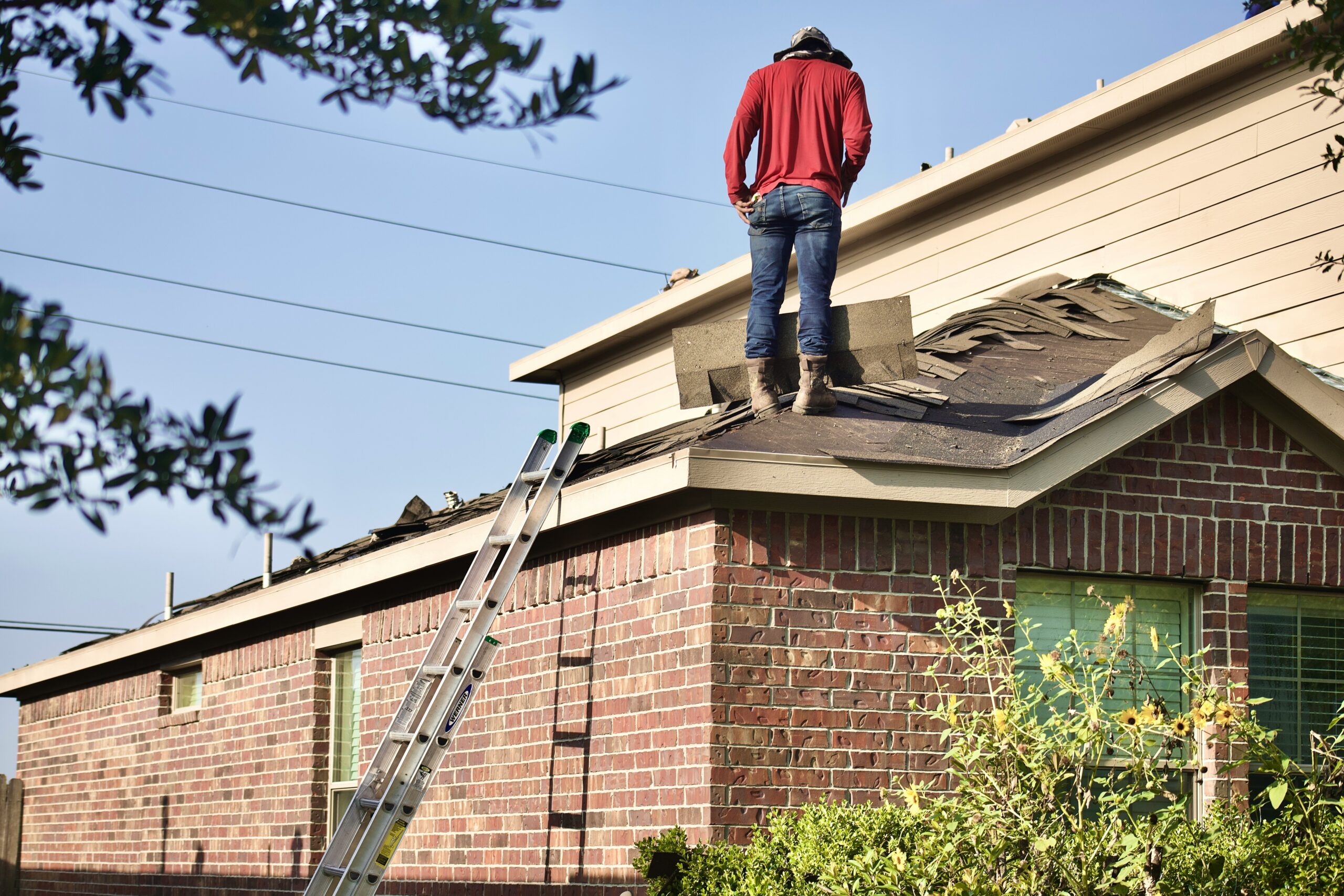 A worker in a red shirt stands on the roof of a brick house, repairing shingles with a ladder leaning against the side of the home.